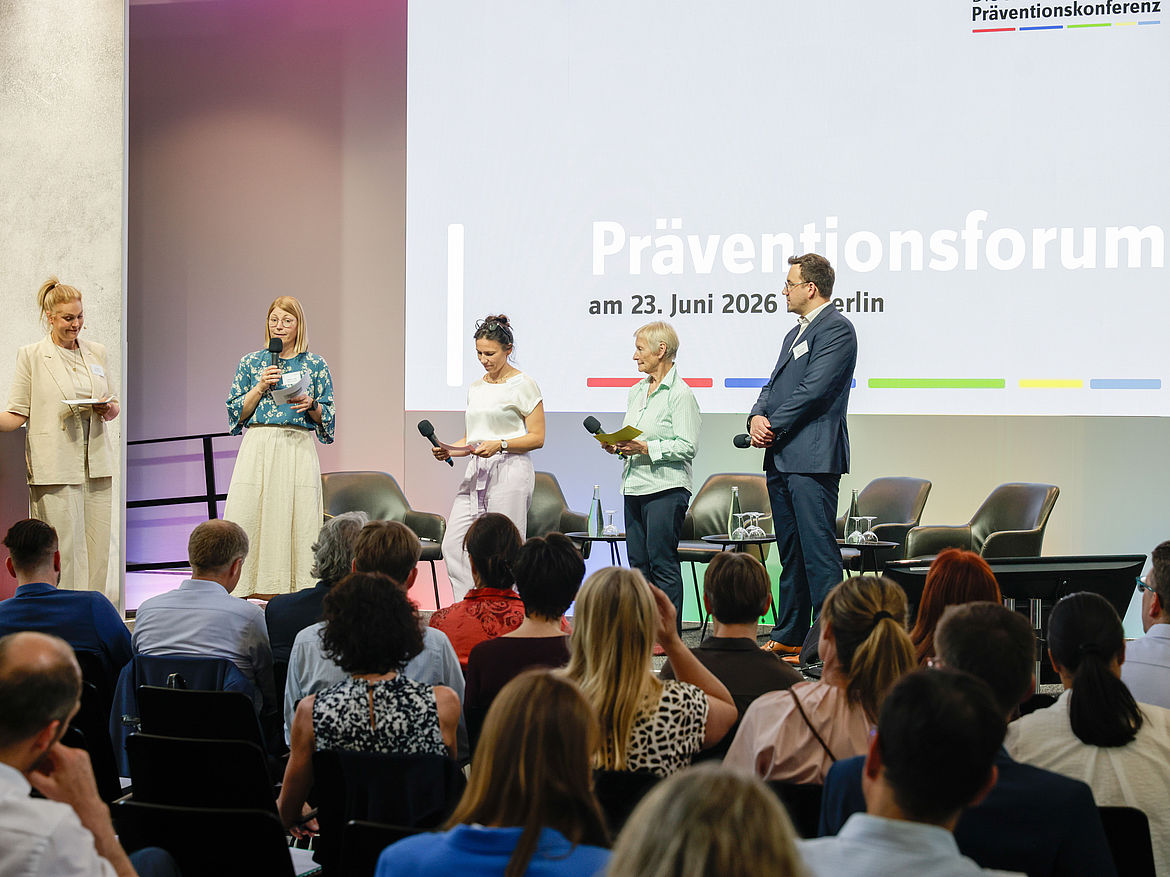 Teilnehmende sitzen in Reihen und blicken auf das Podium, auf dem vier stehende Workshop-Moderierende die Ergebnisse aus ihren jeweiligen Workshops vortragen. 