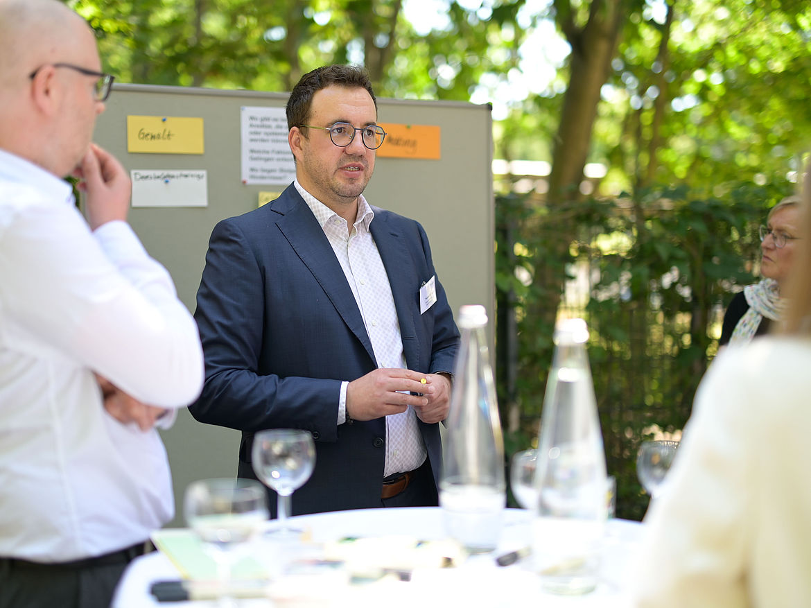 Impression aus Workshop 4 „Gewaltprävention und Mobbing“ Zwei Teilnehmende und ein Moderator stehen im Freien um einen Stehtisch. Der Moderator spricht.