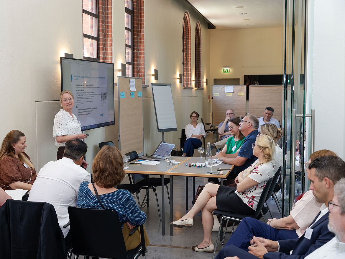 Impression aus Workshop 5 „Führung und mentale Gesundheit“ Eine Workshop-Moderatorin steht vor den Workshop-Teilnehmenden seitlich von einem Tisch. Die Teilnehmenden sitzen kreisförmig um den Tisch.