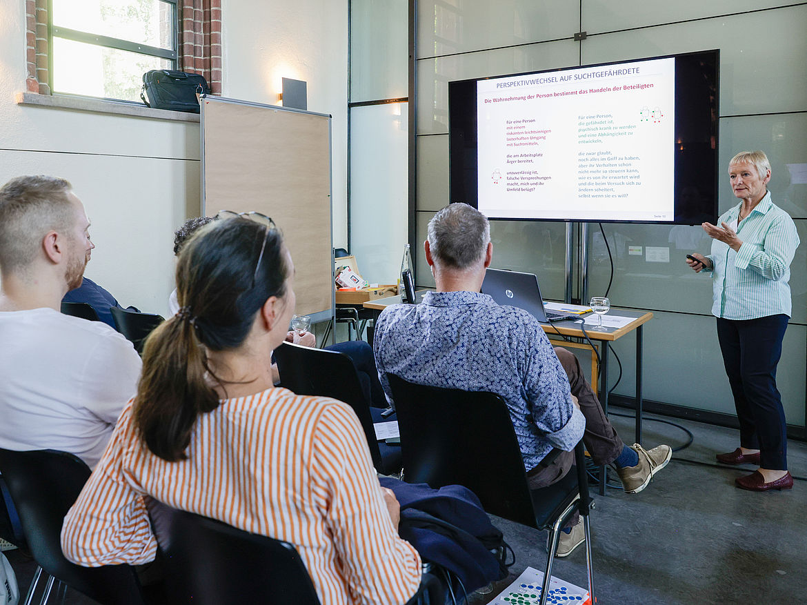 Impression aus Workshop 3 „Suchtprävention und psychische Gesundheit“ Eine Workshop-Moderatorin steht gegenüber den sitzenden Teilnehmenden und führt entlang einer Präsentation in das Thema ein.