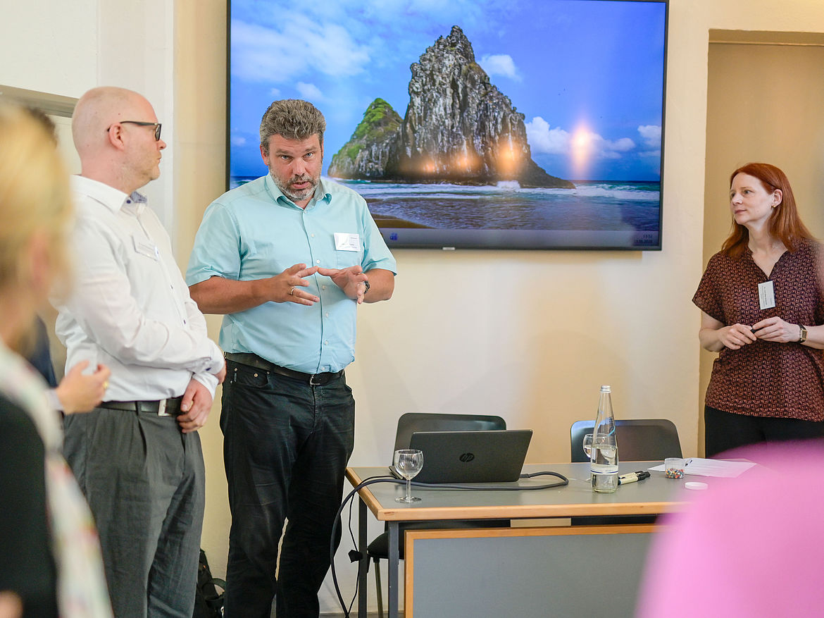 Impression aus Workshop 4 „Gewaltprävention und Mobbing“ Zwei Teilnehmende und zwei Moderierende stehen in einem Seminarraum um einen Tisch, ein Mann erläutert einen Sachverhalt.