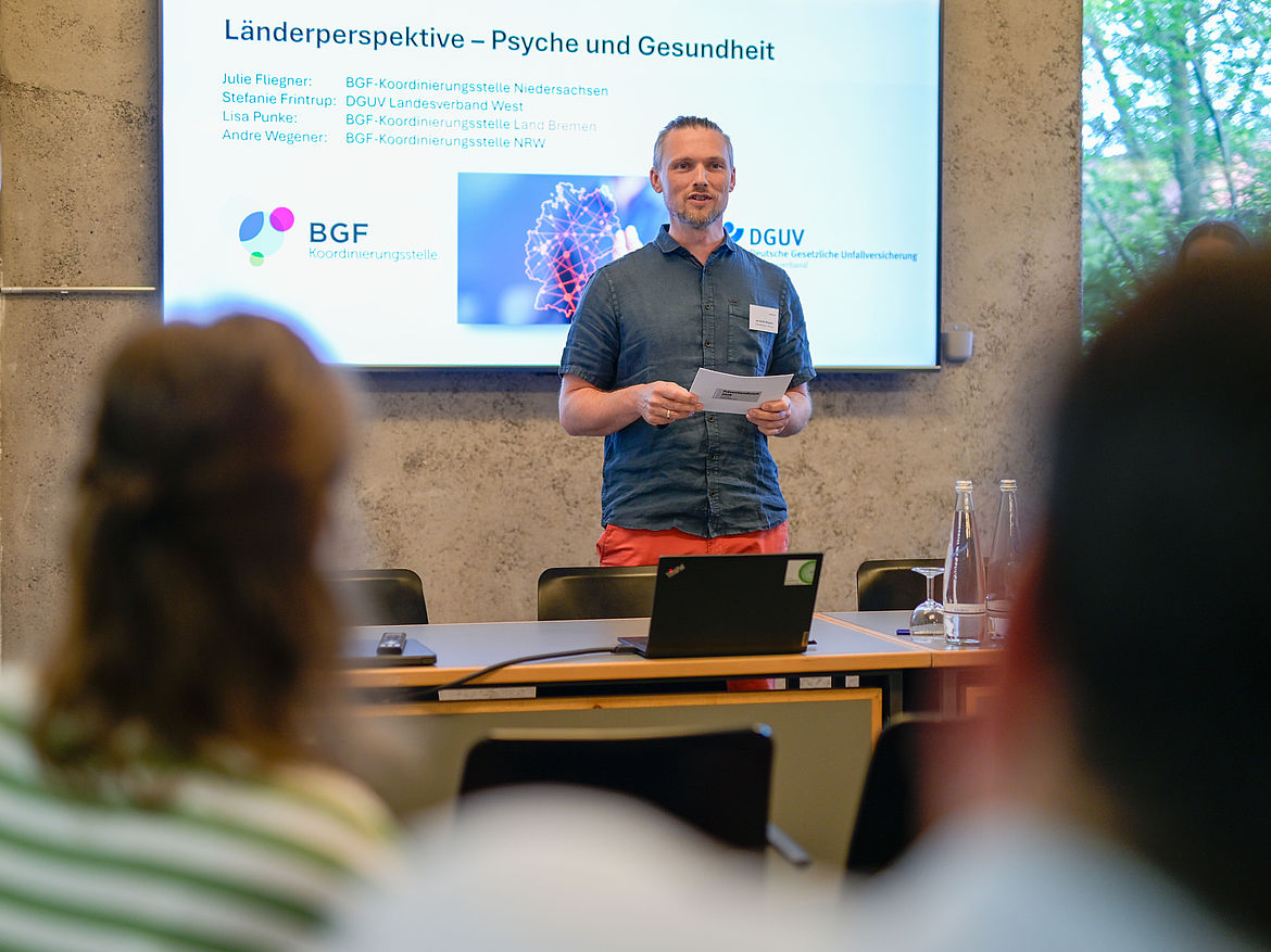Impression aus Workshop 1 „Länderperspektive – Psyche und Gesundheit“ Ein Workshop-Moderator steht an einem Tisch und spricht zu den Teilnehmenden.