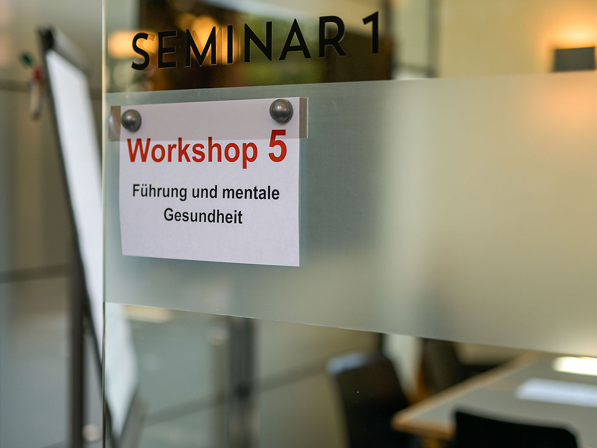 Impression aus Workshop 5 „Führung und mentale Gesundheit“ Am Eingang des Seminarraums hängt ein Schild, auf dem der Workshop 5 „Führung und mentale Gesundheit“ ausgewiesen wird.