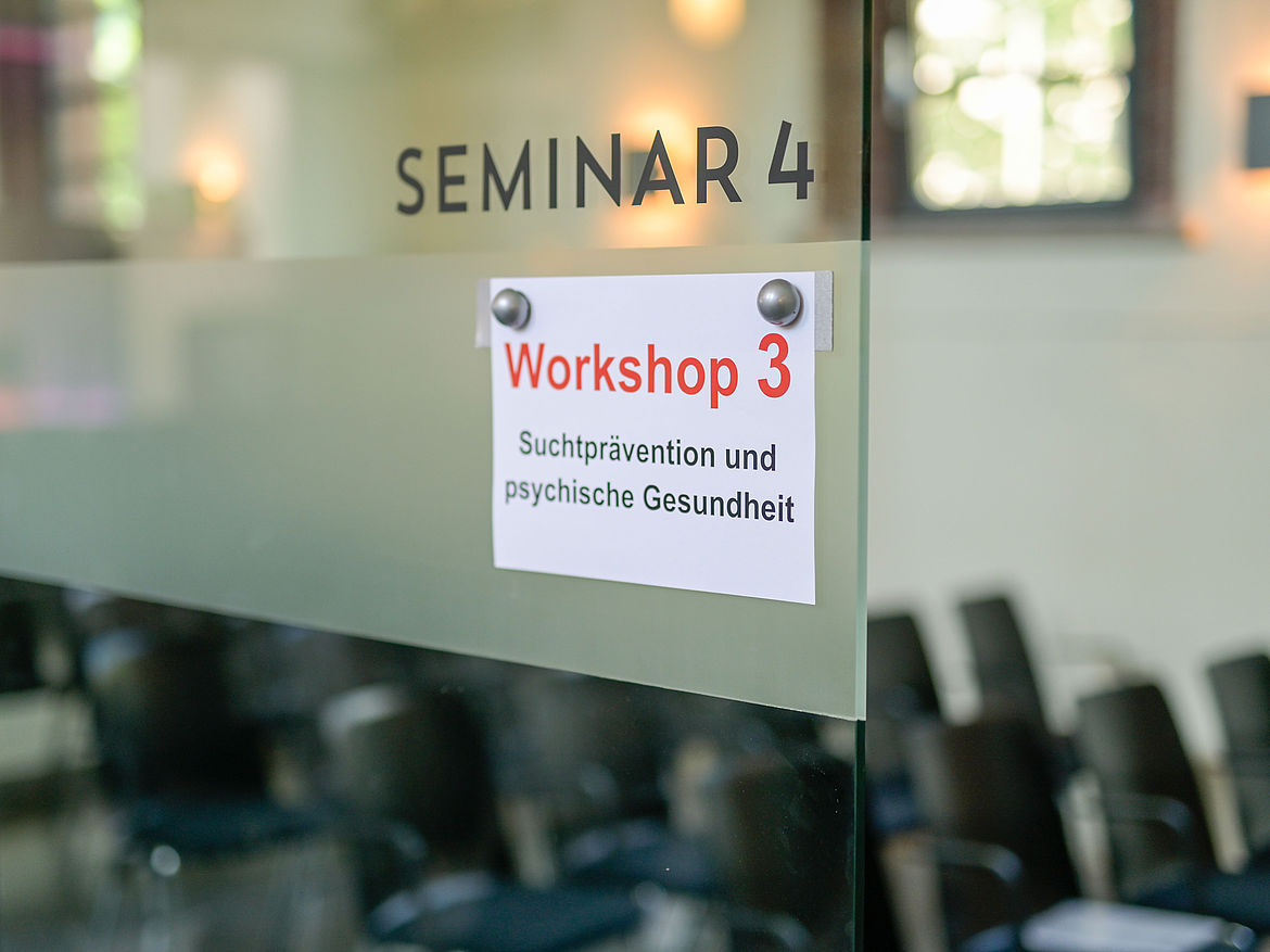 Impression aus Workshop 3 „Suchtprävention und psychische Gesundheit“ Am Eingang des Seminarraums hängt ein Schild, auf dem der Workshop 3 „Suchtprävention und psychische Gesundheit“ ausgewiesen wird.