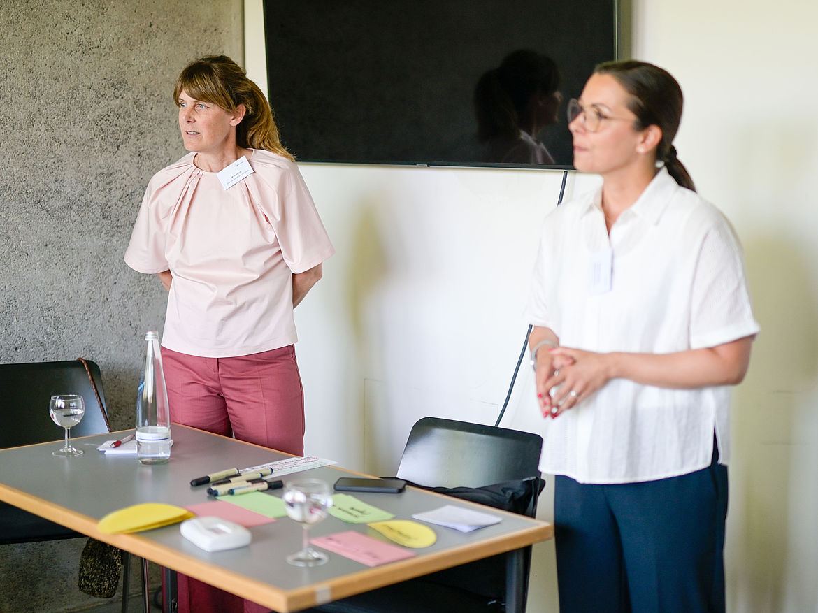 Impression aus Workshop 7 „Offenes Austauschformat – Praxisideen und Impulse“ Zwei Workshop-Moderatorinnen stehen an einem Tisch und schauen zu den Teilnehmenden.