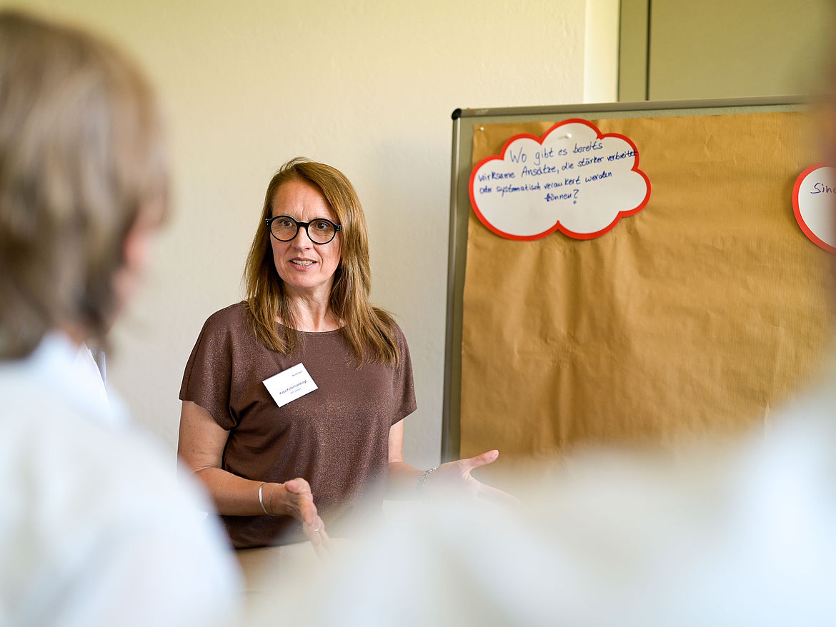Impression aus Workshop 2 „Psychische Gesundheit in kleinen und mittleren Unternehmen (KMU)“ Eine Teilnehmerin steht vor einer Moderationstafel und spricht zu den Teilnehmenden.
