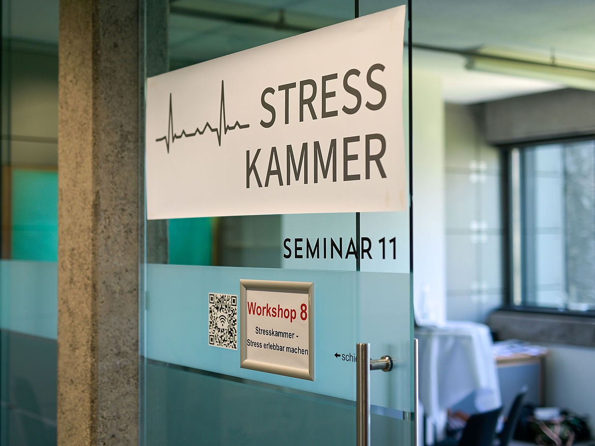 Impression aus Workshop 8 „Stresskammer – Stress erlebbar machen" Am Eingang des Seminarraums hängt ein Schild, auf dem der Workshop 8 „Stresskammer – Stress erlebbar machen“ ausgewiesen wird. Über diesem kleinen Schild hängt ein großes längliches Schild, auf dem das Wort „Stresskammer“ sowie eine Herzfrequenzlinie geschrieben sind.