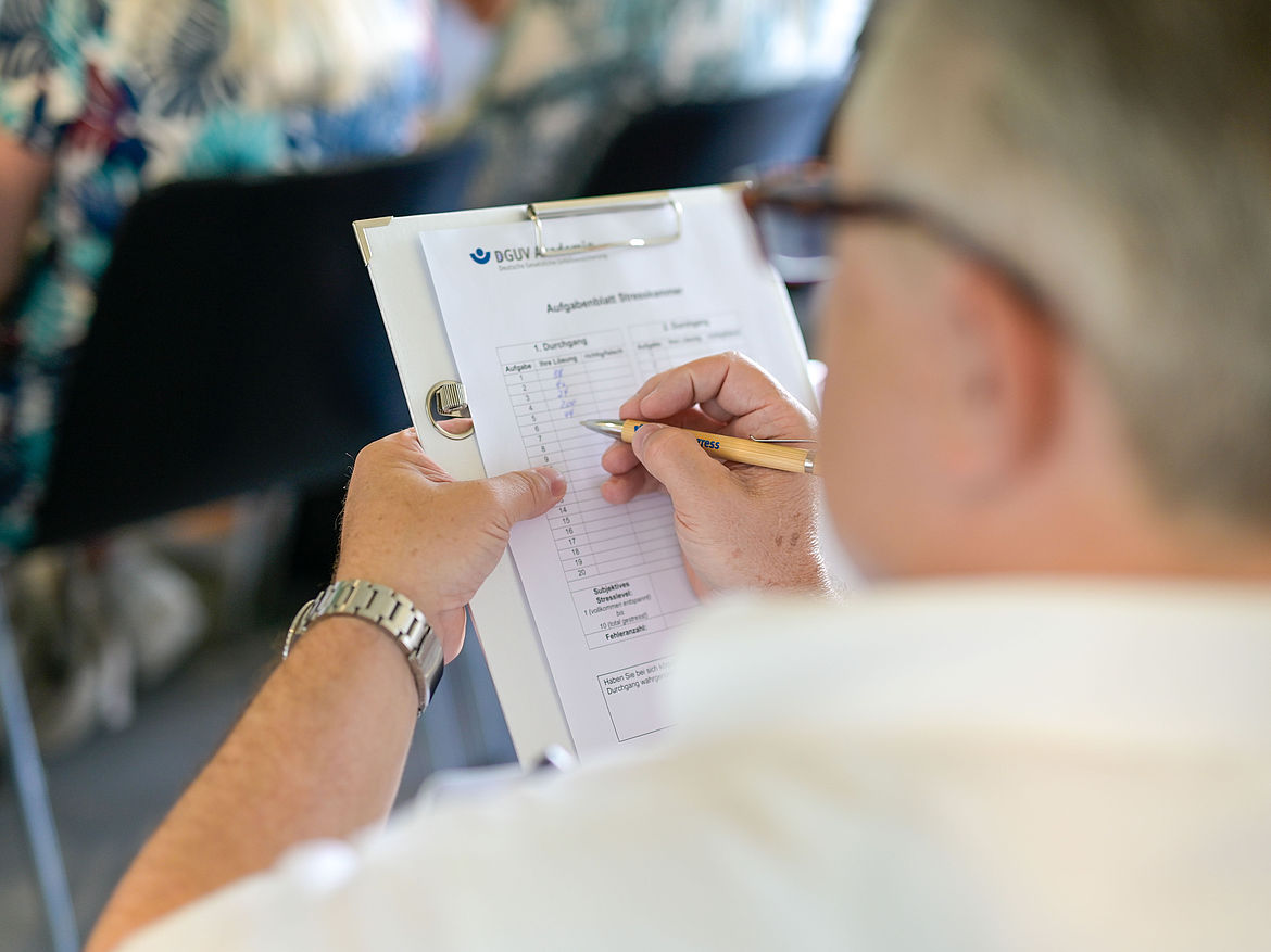 Impression aus Workshop 8 „Stresskammer – Stress erlebbar machen" Ein sitzender Teilnehmender hält ein Klemmbrett mit einer Papiervorlage in der Hand und notiert Werte in eine Tabelle.