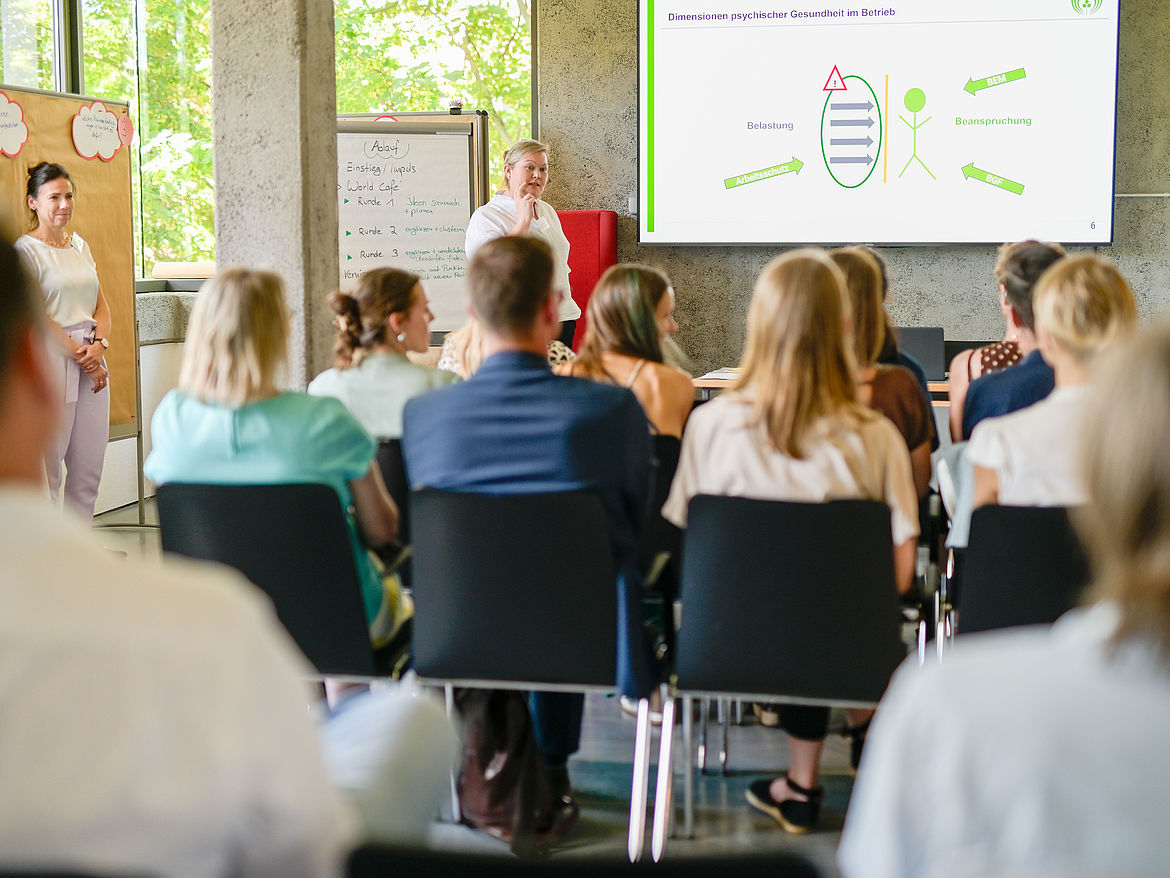 Impression aus Workshop 2 „Psychische Gesundheit in kleinen und mittleren Unternehmen (KMU)“ Die Workshop-Teilnehmenden sitzen in mehreren Reihen und blicken nach vorne auf die Leinwand, vor der die Workshop-Moderatorin steht.