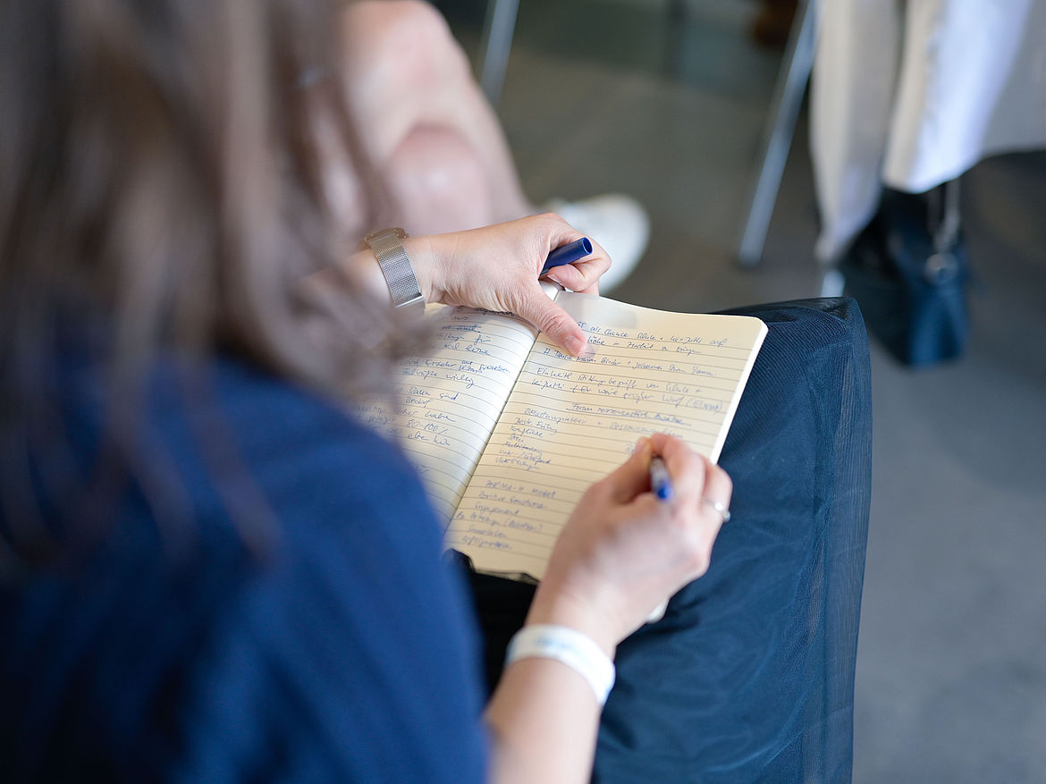 Impression aus Workshop 6 „Ressourcenstärkende Ansätze“ Eine sitzende Teilnehmende schreibt in ein kleines Buch, das auf ihrem Schoß liegt, Notizen.
