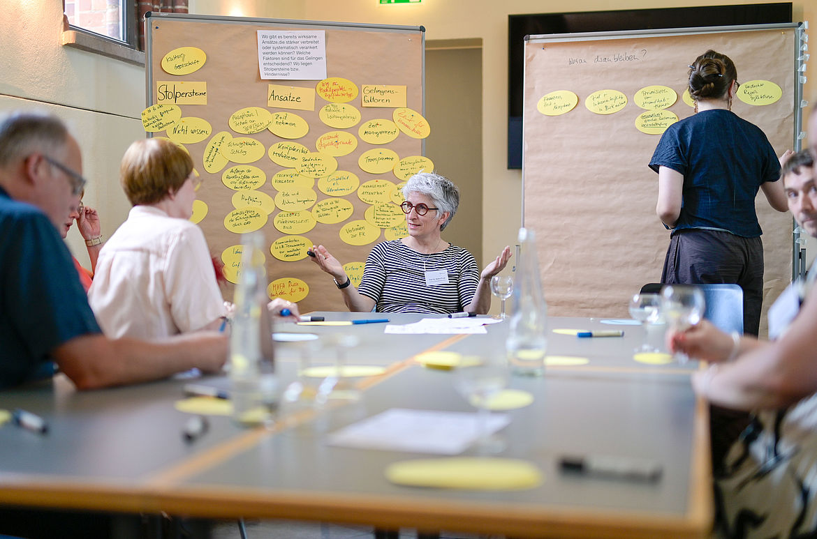 Impression aus Workshop 5 „Führung und mentale Gesundheit“ Eine Workshop-Moderatorin sitzt gemeinsam mit den Teilnehmenden am Tisch. Eine Teilnehmende pinnt eine Antwortkarte an die Moderationstafel.